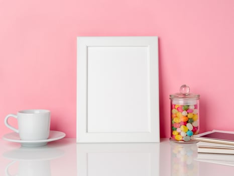 Blank white frame and candys in jar, cup of coffee or tea on a white table against the pink wall with copy space photo