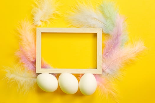 Easter white frame on a bright yellow background of chicken eggs and colored delicate feathers. Spring, religious holiday, Easter decoration, greeting, copy space, mock up photo
