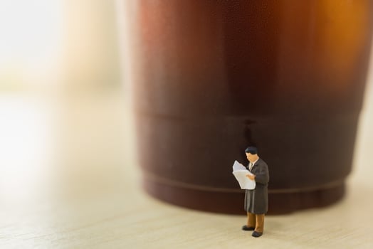 Business, Relaxation Concept. Close up of businessman miniature figure people standing reading newspaper with takeaway plastic cup of iced black coffee on wooden table. photo