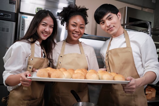 Portrait of three pastry startup partners looking at camera with a cheerful smile and proud with tray of bread in kitchen. A friend and founder of bakery foods and fresh daily bakery small business. photo