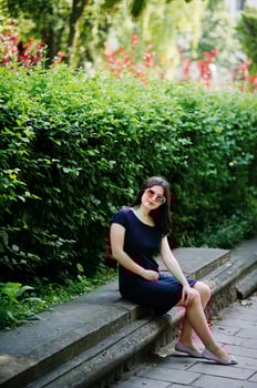 Brunette business girl at black dress on sunglasses sitting at border against bushes, posing at street of city. photo