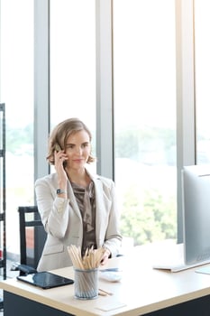 Successful caucasian businesswoman in white suit sitting and talking smartphon at desk in modern office. photo