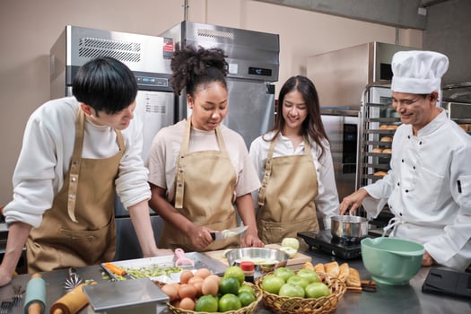 Hobby cuisine course, senior male chef in cook uniform teaches young cooking class students to peel and chop apples, ingredients for pastry foods, fruit pies in restaurant stainless steel kitchen. photo