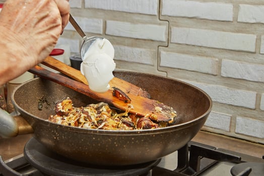 Chef pours fresh cream into pan with fried potatoes, chestnuts, parsley, parmesan and wooden spatulas on gas stove. Step by step recipe photo