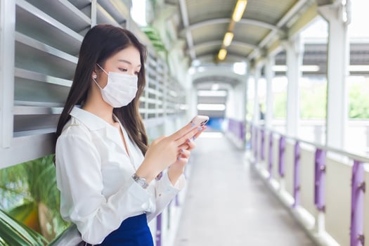 Young Asian beautiful business woman wearing a face mask stands on flyover of skytrain in town while uses her smartphone to send messages to office staff. photo