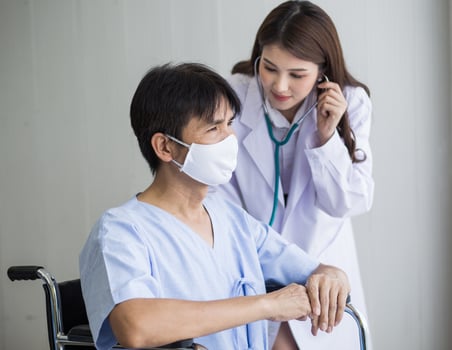 Asian woman doctor use a stethoscope to check the lung rhythm of a male patient who wear face mask while he sit on a wheelchair at hospital. photo