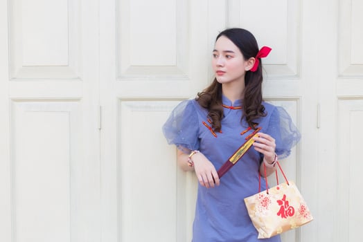 Asian beautiful woman in a blue gray Chinese dress, holding a paper bag with the words happy in Chinese, looking at something and standing on a white wooden door in the background. photo
