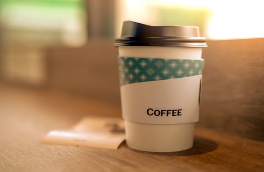 Paper coffee glass in coffee shop on brown wooden table at coffee cafe in the morning with sunlight. Latte, cappuccino, or espresso in paper glass. Disposable paper cup. Green packaging. Hot drink. photo