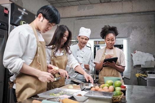 Hobby cuisine course, senior male chef in cook uniform teaches young cooking class students to peel and chop apples, ingredients for pastry foods, fruit pies in restaurant stainless steel kitchen. photo