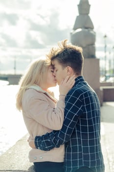 The boy looks tenderly at girl and wants to kiss. A young couple stands embracing. The concept of teenage love and first kiss, sincere feelings of man and woman. The city, the waterfront. Vertical photo