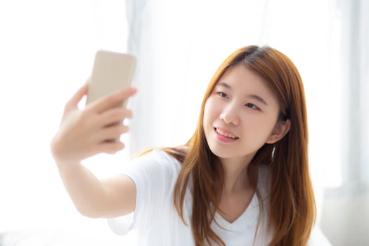 Beautiful portrait young asian woman taking a selfie with smart mobile phone on bedroom, girl is photographing with happy and smile with fun, lifestyle concept. photo