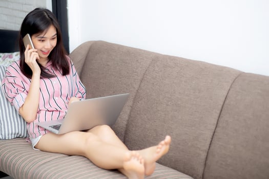 Young woman lying on bed using laptop and talking mobile smart phone on sofa at living room with satisfied for leisure and relax, freelance with girl working notebook, communication concept. photo