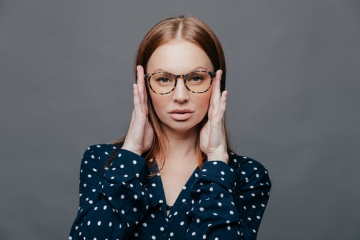 Headshot of serious female teacher keeps hands on head, has self confident facial expression, wears spectacles, polka dot white and black shirt, isolated over grey background. Horizontal shot photo
