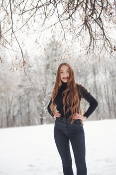 Smiling for the camera. Pretty girl with long hair and in black blouse is in the winter forest photo