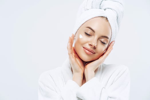 Studio shot of relaxed beautiful woman with closed eyes tilts head, touches skin gently, applies face cream, wears bath towel on head after taking spa procedures, enjoys facial treatments, body care photo