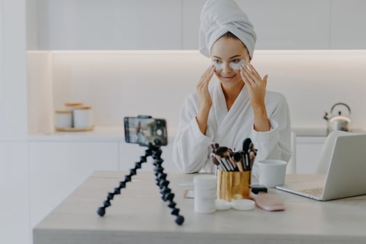 Pretty happy female blogger records streaming video for her blog gives tutorial how to apply beauty patches gives advice about skin care dressed in bathrobe looks at camera poses at table in kitchen photo