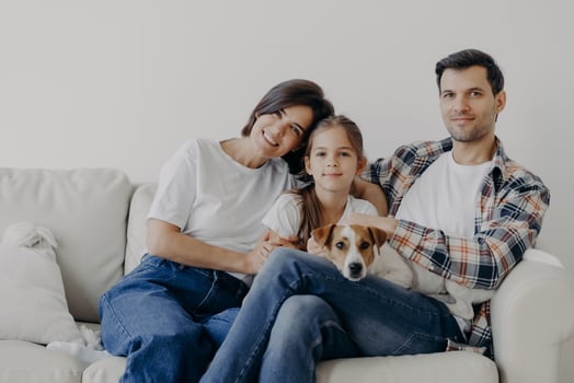 Friendly family pose together at sofa, enjoys domestic atmosphere. Father, mother, their little daughter and pedigree dog spend weekend at home, pose in living room, have happy face expressions photo