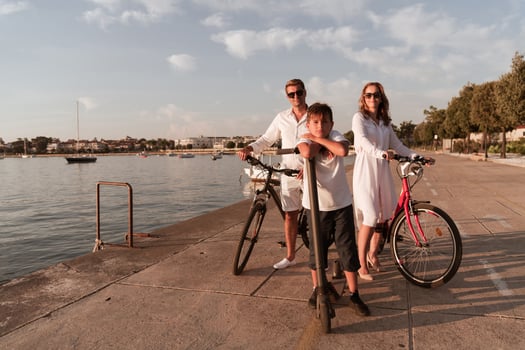 Happy family enjoying a beautiful morning by the sea together, parents riding a bike and their son riding an electric scooter. Selective focus photo