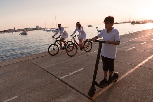 Happy family enjoying a beautiful morning by the sea together, parents riding a bike and their son riding an electric scooter. Selective focus photo