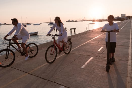 Happy family enjoying a beautiful morning by the sea together, parents riding a bike and their son riding an electric scooter. Selective focus photo