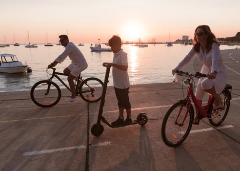 Happy family enjoying a beautiful morning by the sea together, parents riding a bike and their son riding an electric scooter. Selective focus photo