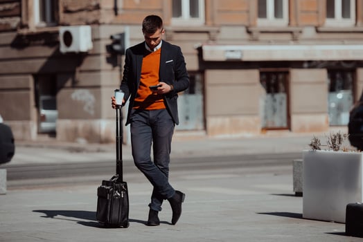 Going to airport terminal. Confident businessman traveler walking on city streets and pulling his suitcase drinking coffee and speaking on smartphone photo