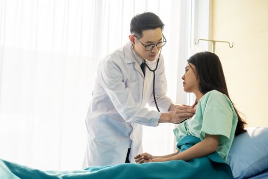 Asian male doctor wearing stethoscope on ear and taking the heartbeat of a female patient sitting on a hospital bed photo