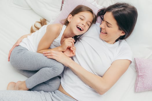 Photo of adorable small child and her mother have fun together in bed, tickle each other, smile joyfully, play after good sleep, have good relationships. People, family, motherhood and childhood