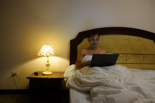 A smiling young man is sitting in bed in the evening working on a laptop, freelancer at home. photo