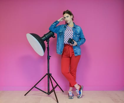 Young woman leaning against studio flashlight and holding camera over a pink background photo
