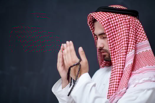 arabian man making traditional prayer to God, keeps hands in praying gesture in front of black chalkboard photo