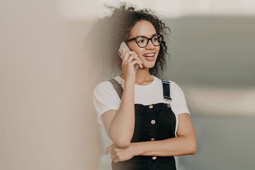 Smiling young curly woman with gentle smile, talks on mobile phone, has friendly conversation, wears casual white t shirt and overalls, focused aside. Leisure time, lifestyle, technology concept photo