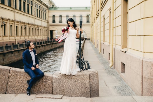 Handsome bridegroom sits on bridge looks at his beautiful bride who stands up, says compliments to her, enjoy togetherness, have unforgettable moments together, Romantic people and wedding concept photo