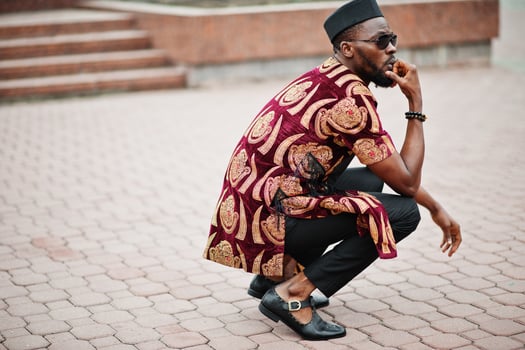 African stylish and handsome man in traditional outfit and black cap squatted outdoor. photo