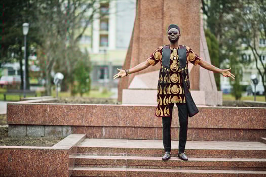 African stylish and handsome man in traditional outfit, sunglasses and black cap standing outdoor against monument. photo