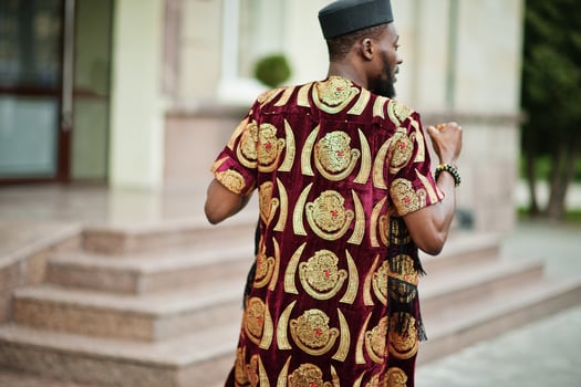 Back of african stylish and handsome man in traditional outfit and black cap standing outdoor. photo