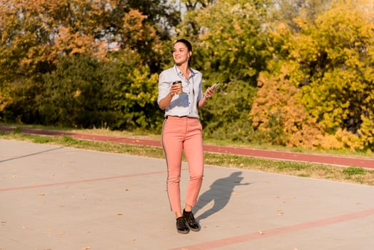 Happy young woman with coffee listening music on smartphone and walking on promenade photo