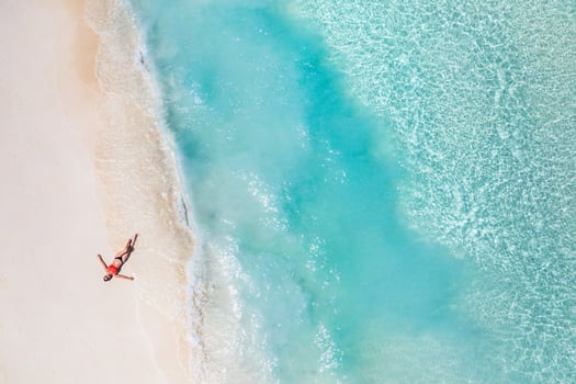 Top aerial view from drone young woman in a bikini lying on the back on the white sand near the waves of blue sea. Beautiful woman tans on sandbank surrounded by turquoise ocean from above photo