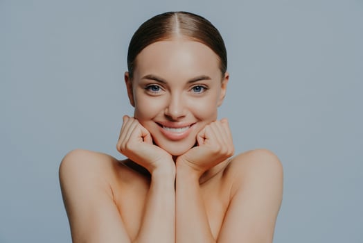 Women, skin care and tenderness concept. Young female takes care of skin and body health, keeps hands under chin and smiles gently, wears makeup, has combed hair, delighted with skin care product photo