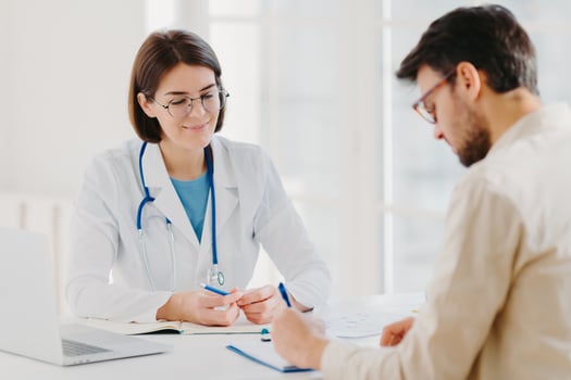 Male patient writes down application form, gets consultancy and advice about home treatment from doctor, use laptop and medical document at desk, pose at office together. Man doctors appointment photo