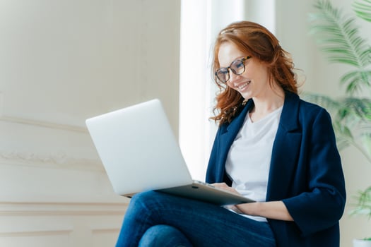 Professional female employer enjoys working process, sits crossed legs with laptop device, chats online, has positive look, has red curly hair, wears stylish elegant clothes. People and technology photo