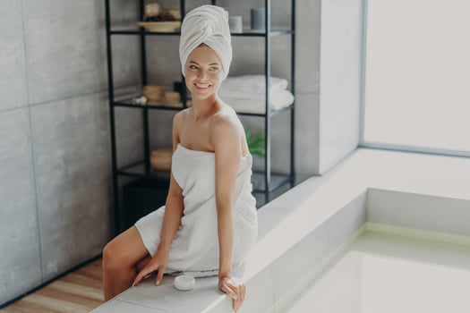 Beauty, grooming and hygiene concept. Relaxed beautiful European woman with healthy skin smiles and looks away, wrapped in bath towel, uses cosmetic cream, enjoys beauty routine, takes shower photo