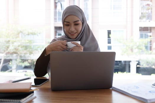 Asian Muslim hijab woman holding coffee cup looking at computer smiling happily photo