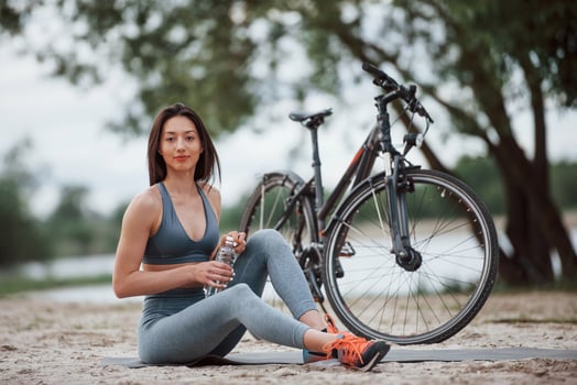 Do not forget take water for your exersices. Female cyclist with good body shape sitting near her bike on beach at daytime photo