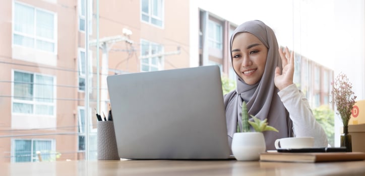 smiling beautiful muslim businesswoman using a laptop for online video meeting sitting at the desk in modern office space, looks at the webcam and waving hello photo