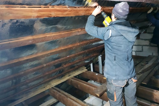 The wizard uses a cordless drill and screw the screws into boards that hold foil film and mineral wool to heal the roof of the house photo