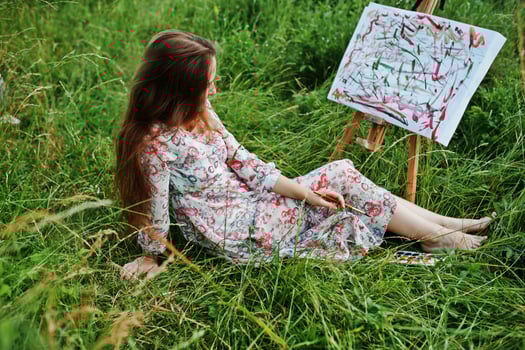 Portrait of a gorgeous happy young woman in beautiful dress sitting on the grass and painting on paper with watercolors. photo
