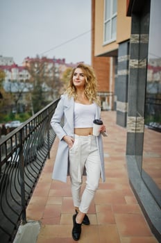 Stylish curly blonde model girl wear on white with cup of coffee at hand posing against large window. photo
