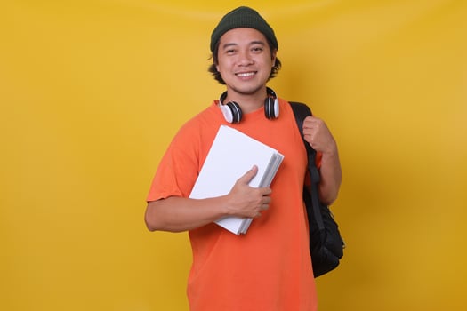 Asian young man in casual style with backpack and headphone bringing books isolated on yellow background. School college university concept. photo