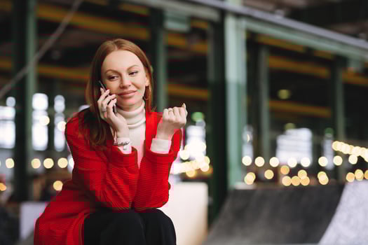 People, technology and lifestyle concept. Positive Caucasian woman has telephone conversation, talks to friends via cellular, wears red coat and black trousers, poses outside with free space photo